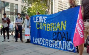 A banner reading "Leave Cumbrian Coal Underground" is held by 5 protestors outside the Ministry of Levelling up in London, on a sunny day.